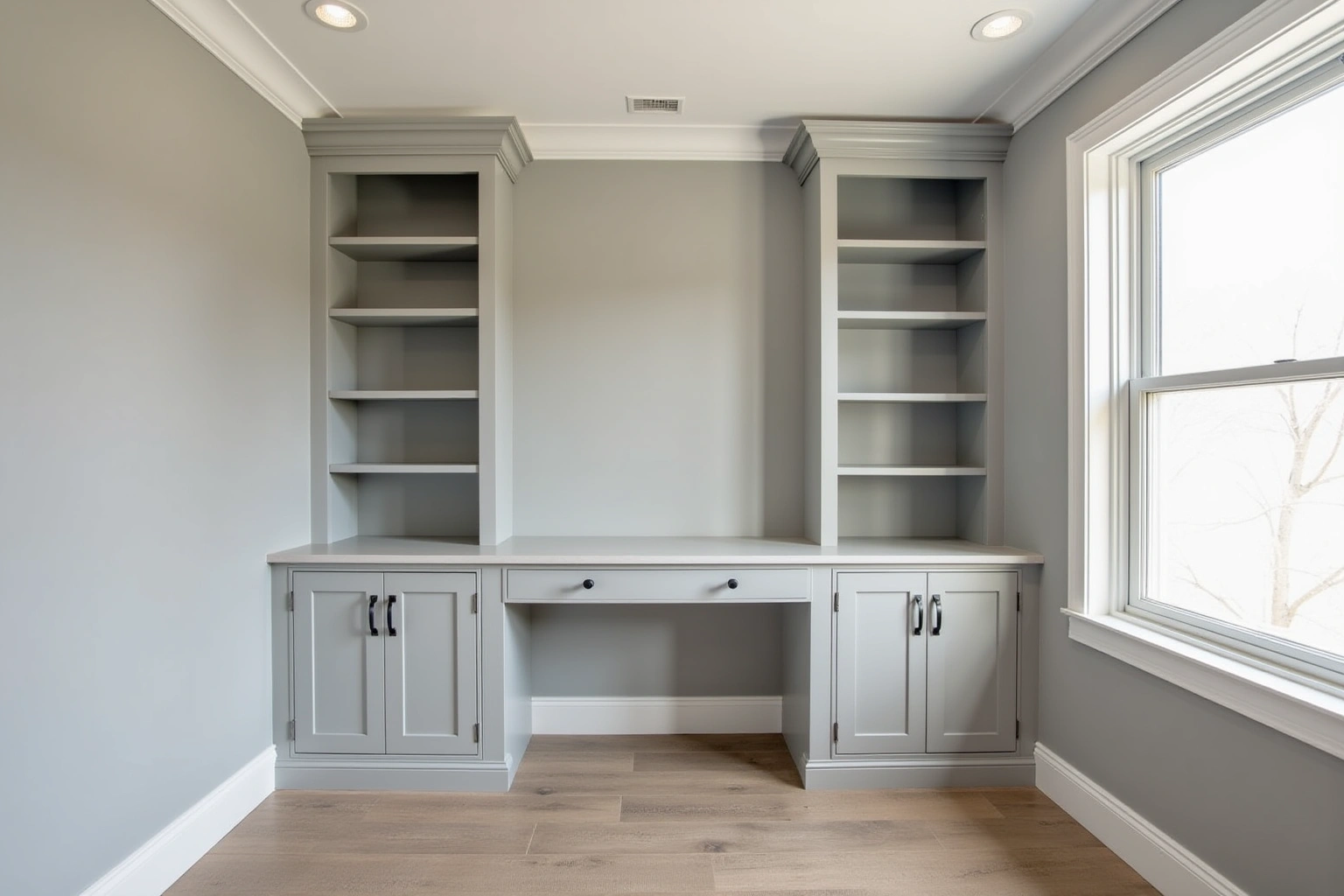 Grey painted home office built-ins with open shelving towers, a built-in desk with a drawer, crown molding and a window.